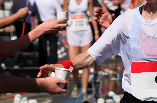Runner grabbing or drinking water from a cup at a race aid station, mid-stride, crowd blurred behind
